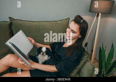 femme avec des patchs sous les yeux lisent le livre sur un canapé vert avec un chien menton japonais noir et blanc dans un intérieur confortable avec un éclairage doux et une plante verte Banque D'Images