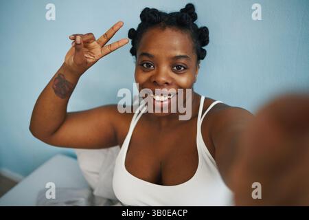 Portrait de femme afro-américaine souriante prenant selfie avec signe de paix tout en étant assise sur le lit portant un haut blanc et maquillage naturel profitant de sa journée Banque D'Images