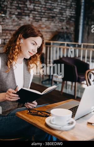 Une jeune femme aux cheveux roux bouclés prend des notes alors qu'elle participe à une réunion en ligne dans un café. Réseautage moderne, présence virtuelle, engagement numérique Banque D'Images