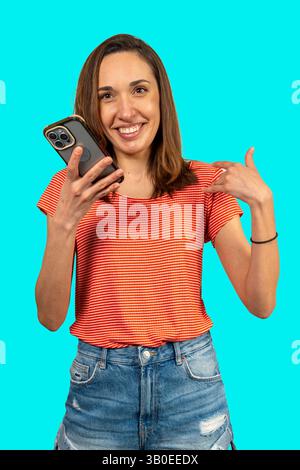 Jeune femme pointant son smartphone avec un grand sourire sur fond turquoise Banque D'Images