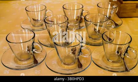 Un ensemble de huit tasses en verre transparent avec des soucoupes disposées soigneusement sur une nappe de couleur claire. Chaque tasse a une petite cuillère placée sur sa soucoupe, créant Banque D'Images
