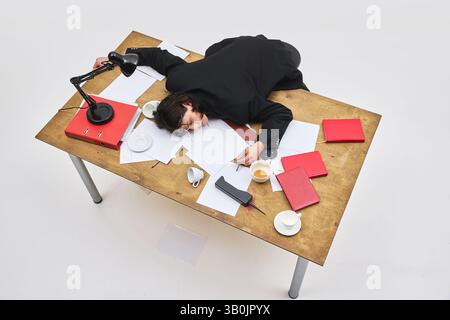 Vue aérienne d'un employé épuisé s'est effondré sur un bureau encombré avec du café renversé et des documents éparpillés. Banque D'Images