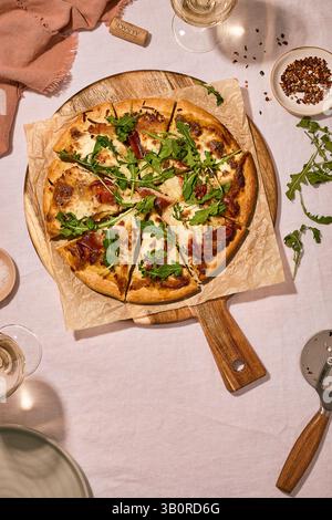 Pizza artisanale avec prosciutto et roquette sur pagaie en bois, accompagnée de vin blanc, liège de vin et mélange d'épices dans un cadre élégant. Banque D'Images