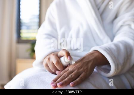 La femme pompe la lotion du distributeur blanc sur sa main dans un peignoir près de la plante en pot Banque D'Images