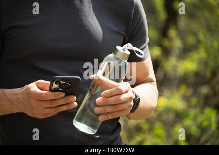Homme de sport tenant la bouteille d'eau et vérifier les applications sur le terrain de sport extérieur. Concept équilibre de l'eau pour la saison chaude pour l'entraînement. Gros plan. Banque D'Images