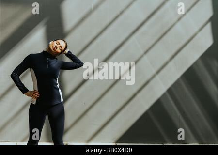 Une femme gracieuse effectue un étirement du cou, profitant de la lumière douce du soleil filtrant à travers les lignes architecturales dans une salle de sport moderne. Banque D'Images