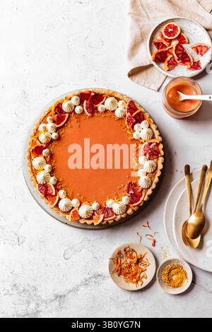 Une tarte à l'orange garnie de crème fouettée et de tranches d'orange sanguine, entourée de divers ustensiles de cuisine et ingrédients sur une surface blanche. Banque D'Images