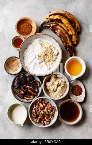 Assortiment d'ingrédients pour une recette comprenant des bananes mûres, diverses noix, de la farine et des épices disposées sur une surface texturée. Banque D'Images
