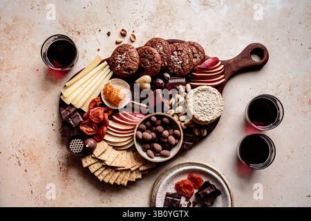 Un assortiment de collations comprenant du fromage, des biscuits, des fruits secs et des chocolats disposés sur une planche de bois, accompagnés de verres de vin rouge. Banque D'Images