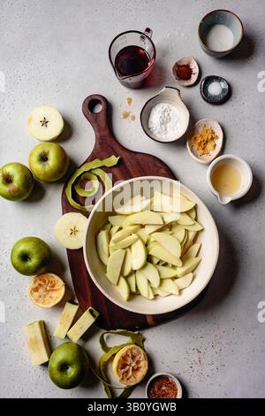 Un assortiment de pommes vertes, tranchées et préparées pour la cuisson, ainsi que divers ingrédients comme la farine, les épices et le citron sur une planche à découper en bois. Banque D'Images
