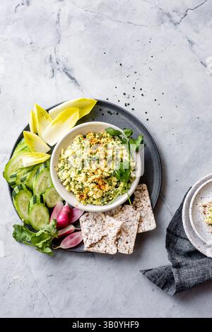 Une assiette avec un bol de salade crémeuse garnie d'herbes, entourée de tranches de concombre, de feuilles d'endive, de radis et de gâteaux de riz. Banque D'Images