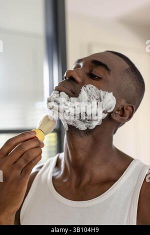 Homme afro-américain étalant la mousse à raser sur le visage dans la salle de bain, avec brosse à raser Banque D'Images