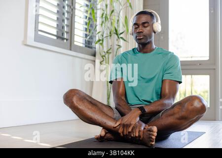 Homme afro-américain méditant assis les jambes croisées dans le coin de méditation avec tapis de yoga, écouteurs Banque D'Images