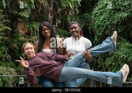 Deux jeunes femmes heureuses tenant et soulevant un troisième ami faisant signe de paix devant un mur vert couvert de végétation pendant une journée ensoleillée, ayant f Banque D'Images