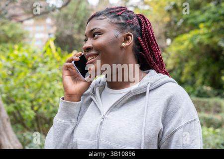 Heureuse jeune femme noire souriante tout en utilisant son téléphone portable dans un parc, profitant d'une conversation par une journée ensoleillée, portant un sweat à capuche gris et des tresses rouges Banque D'Images