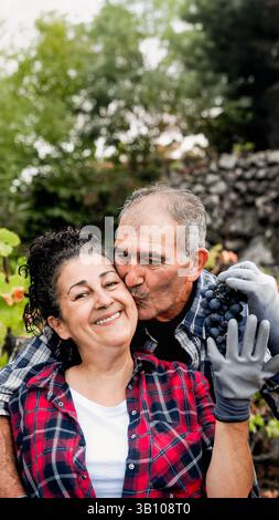 Agriculture, fermier couple senior s'amusent à tenir un bouquet de raisins. Les gens heureux s'embrassent et s'embrassent à la ferme pendant la saison des récoltes. Petite entreprise et produ vin Banque D'Images