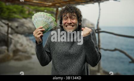 Homme hispanique tenant joyeusement des roubles russes sur une plage de bord de mer, exprimant l'excitation à l'extérieur dans un pull. Banque D'Images