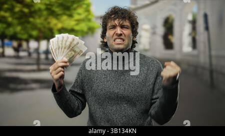 Homme hispanique tenant des roubles russes à l'extérieur sur la rue de la ville, exprimant l'excitation, portant un pull décontracté, avec des arbres et l'amélioration de fond urbain Banque D'Images