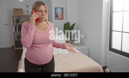 Femme enceinte aux cheveux blonds se tient dans une chambre à coucher parlant au téléphone, attendant un bébé, entourée d'un intérieur confortable, exprimant son inquiétude. Banque D'Images