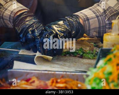 Gros plan des mains dans des gants bleus et des manches à carreaux préparant un sandwich avec des ingrédients frais Banque D'Images