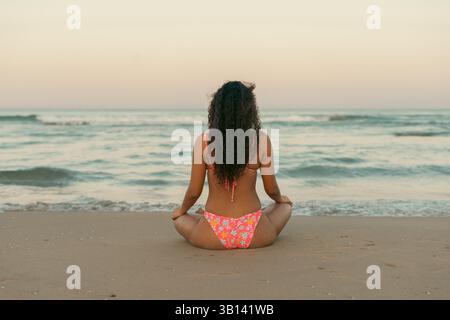 Femme aux cheveux bouclés assis les jambes croisées sur une plage de sable, méditant alors que le soleil se couche sur l'océan calme. Le cadre serein met l'accent sur la détente et Banque D'Images