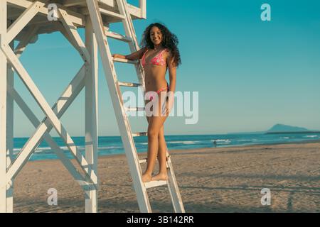 Femme dans un maillot de bain coloré se tient sur une échelle de tour de sauveteur à une plage de sable, avec l'océan et un ciel bleu clair en arrière-plan, transportant un s. Banque D'Images
