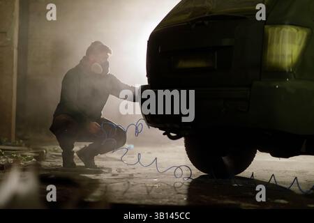 Un mécanicien se concentre sur la réparation de l'arrière d'un véhicule dans un garage faiblement éclairé. L'atmosphère est remplie de poussière et la lueur de l'equipme environnant Banque D'Images