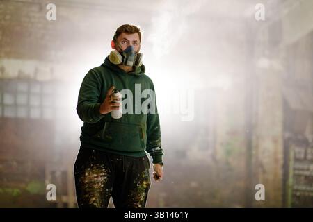 Un artiste portant un respirateur tient une bombe de peinture en aérosol, debout dans un entrepôt désert. La zone est marquée avec de la poussière et des éclaboussures de peinture, highlighti Banque D'Images