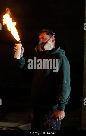 Un créateur dans un studio sombre tient une bombe aérosol qui produit des flammes, portant un masque pour la sécurité. Cette activité unique mêle art et feu, mettant en valeur le créa Banque D'Images