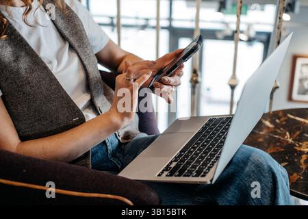 Gros plan des mains d'une jeune femme alors qu'elle interagit avec son smartphone tout en travaillant sur un ordinateur portable. Habitudes de travail multi plates-formes, intégration de périphériques Banque D'Images