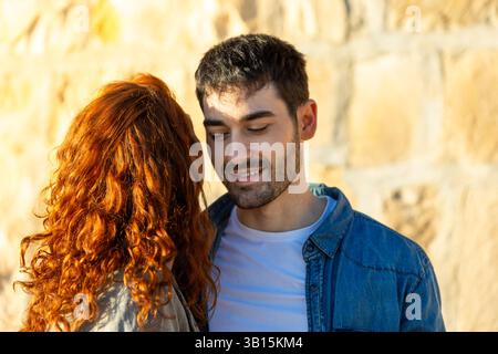 Gros plan d'un jeune couple partageant un moment tendre, avec la femme penchée à proximité et l'homme souriant doucement, debout contre un mur de pierre chaude bac Banque D'Images