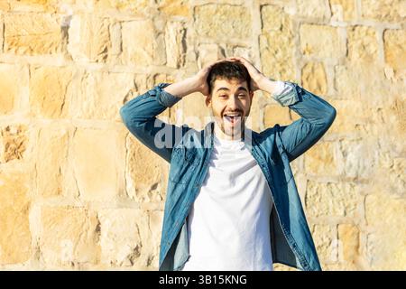 Jeune homme en chemise en denim et t-shirt blanc exprimant l'excitation et la surprise avec un large sourire et les mains sur la tête contre un fond de mur de pierre brillant Banque D'Images