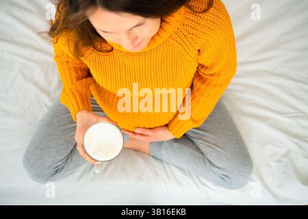 Vue de haut en bas d'une femme enceinte en position de lotus tenant un latte, contemplant les choix alimentaires pendant la grossesse. Moment chaleureux de soins personnels et de conscience. Banque D'Images