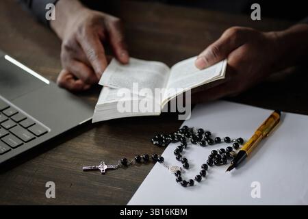 Gros plan moyen de mains de pasteurs méconnaissables tenant la Bible ouverte. Il est assis à un bureau en bois avec des ordinateurs portables, stylo vintage et chapelet avec des perles noires et croix Banque D'Images