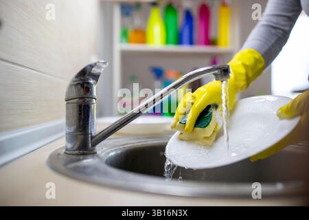 Femme portant des gants jaunes frotte la plaque dans l'évier de cuisine rempli d'eau, entourée de fournitures de nettoyage colorées sur les étagères en arrière-plan Banque D'Images