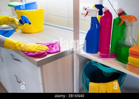 Personne portant des gants jaunes essuyant le comptoir de cuisine avec un chiffon tout en étant entourée de fournitures de nettoyage colorées organisées à proximité Banque D'Images
