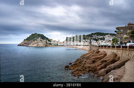 Une vue panoramique sur la côte avec des rivages rocheux, une plage de sable et une charmante ville nichée sur la colline. Le ciel est couvert, créant une atmosphère sombre Banque D'Images