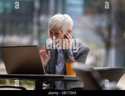 Gestionnaire plus âgée assise à l'extérieur pour prendre un café et passer un appel vidéo sur ordinateur portable Banque D'Images