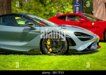 Une élégante supercar grise McLaren 765LT stationnée sur une zone herbeuse, avec une Ferrari rouge en arrière-plan, lors d'un salon automobile en plein air de jour. Banque D'Images
