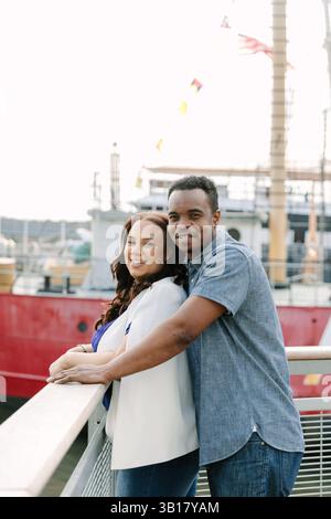 Joyeux couple pose près de la balustrade près de l'eau, avec un navire historique amarré derrière eux par un après-midi lumineux. Banque D'Images