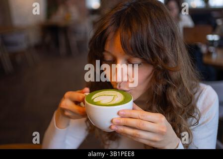 Jeune femme aux longs cheveux bruns savoure une tasse de latte matcha dans un café confortable, soufflant doucement sur la surface verte vibrante ornée d'art latte délicat Banque D'Images