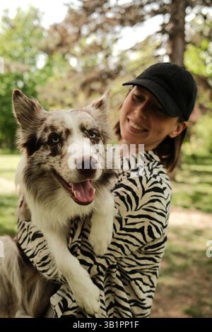 Heureuse jeune femme tenant un adorable chien rouge merle Border Collie dans ses bras, tous deux souriants dans un parc ensoleillé. Moment lumineux et joyeux plein d'amour et Banque D'Images