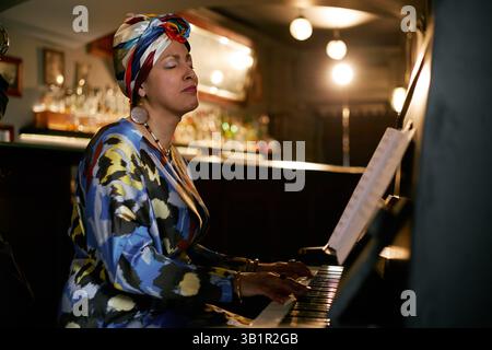 Vue latérale d'une artiste passionnée portant un élégant couvre-tête jouant du piano tout en jouant de la musique live dans un club de jazz remplissant la salle avec une musique enchanteresse Banque D'Images