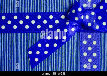 Gros plan d'un ruban cadeau bleu avec des pois blancs attachés dans un noeud, placé sur un fond rayé rouge et rose texturé. Banque D'Images