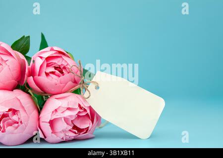 Un bouquet serré de pivoines roses fraîches avec une étiquette cadeau en papier vierge, fond bleu doux et audacieux avec espace de copie Banque D'Images