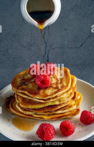une pile de crêpes à la banane dorée sur une assiette blanche, surmontée de framboises fraîches, comme le sirop d'érable est versé sur eux Banque D'Images