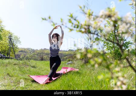 Femme effectuant une pose de fente basse sur un tapis rose dans un champ vert, bras gracieusement tendus au-dessus de la tête, vue latérale, dessus rayé, arbres en fleurs. Banque D'Images