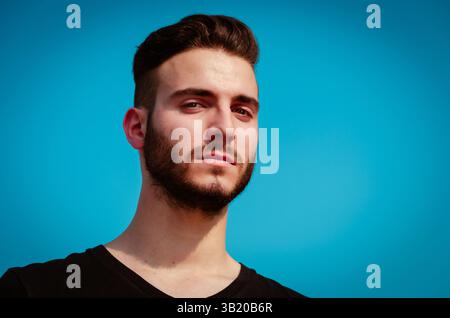 Un jeune homme se tient confiant avec une expression sérieuse. Il a les cheveux courts et une barbe bien soignée, portant une simple chemise noire tandis que le ciel brille derrière lui. Banque D'Images