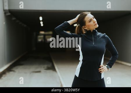 Une jeune femme pose en toute confiance avec une main sur son cou tout en se tenant debout dans un cadre urbain moderne, prête pour sa séance d'entraînement. Banque D'Images