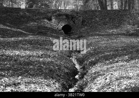 Le trou noir d'un vieux drain en brique avec un petit ruisseau divisant le ravin en deux dans le style noir et blanc Banque D'Images